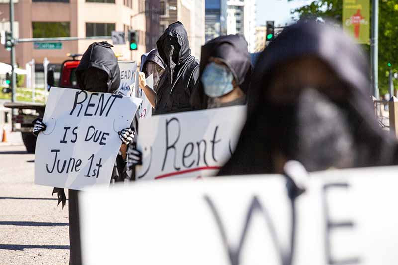 Protestors, some with their faces fully covered, stand in a city street holding signs about rent